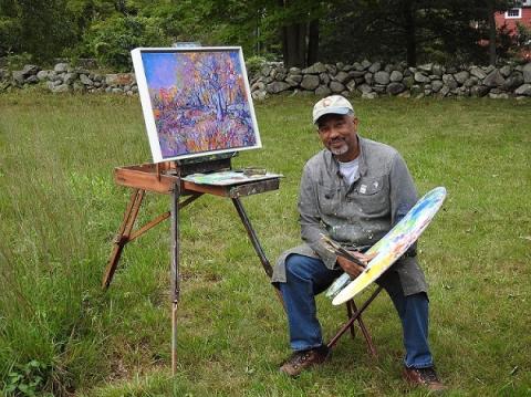 Black man wearing a baseball cap, seated before an easel and painting outside and holding a large palette and paintbrush
