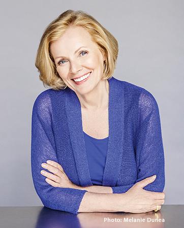 White woman with strawberry blonde hair smiling and tilting her head to the right while leaning crossed arms on table