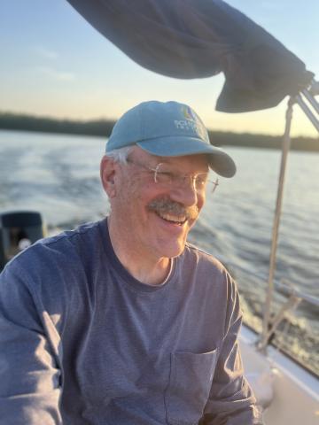 White man with glasses and mustache wearing a light blue baseball cap with ocean view behind him