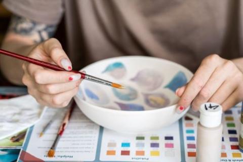 Image of hands holding a paint brush as they paint a bowl in colorful geometric shapes