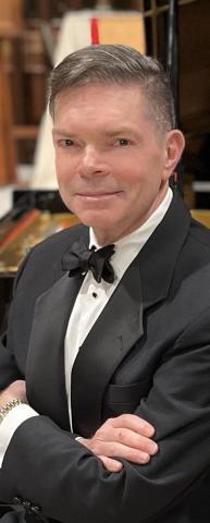 White man with short brown hair wearing a tuxedo, looking at camera with arms crossed in front of himself