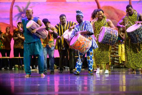 Black musicians standing with drums and performing against a pink background while wearing colorful traditional West African clothing