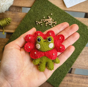 photo of a hand holding a felt frog wearing a flower around his head