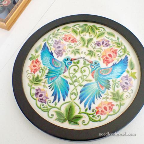 Two embroidered blue hummingbirds face each other. There is delicate filigree surrounding them.