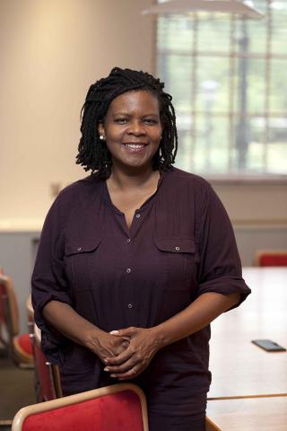 3/4 length photo of Black woman with shoulder length hair and hands folded in front of her smiling at camera