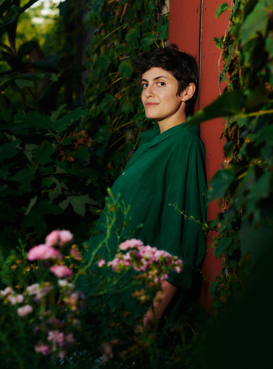 Woman with short brown hair standing against wall with plants around her and looking to her left