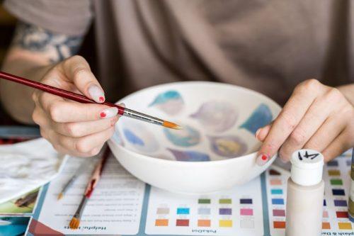 Image of hands holding a paint brush as they paint a bowl in colorful geometric shapes